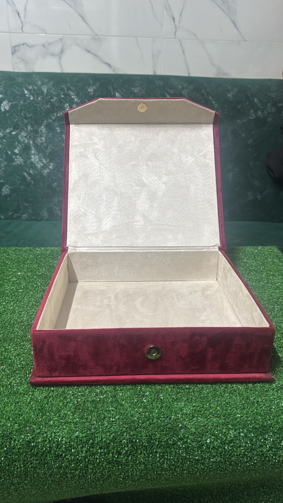 Coffret vide en velours rouge avec intérieur doublé velours, posé sur fond vert.